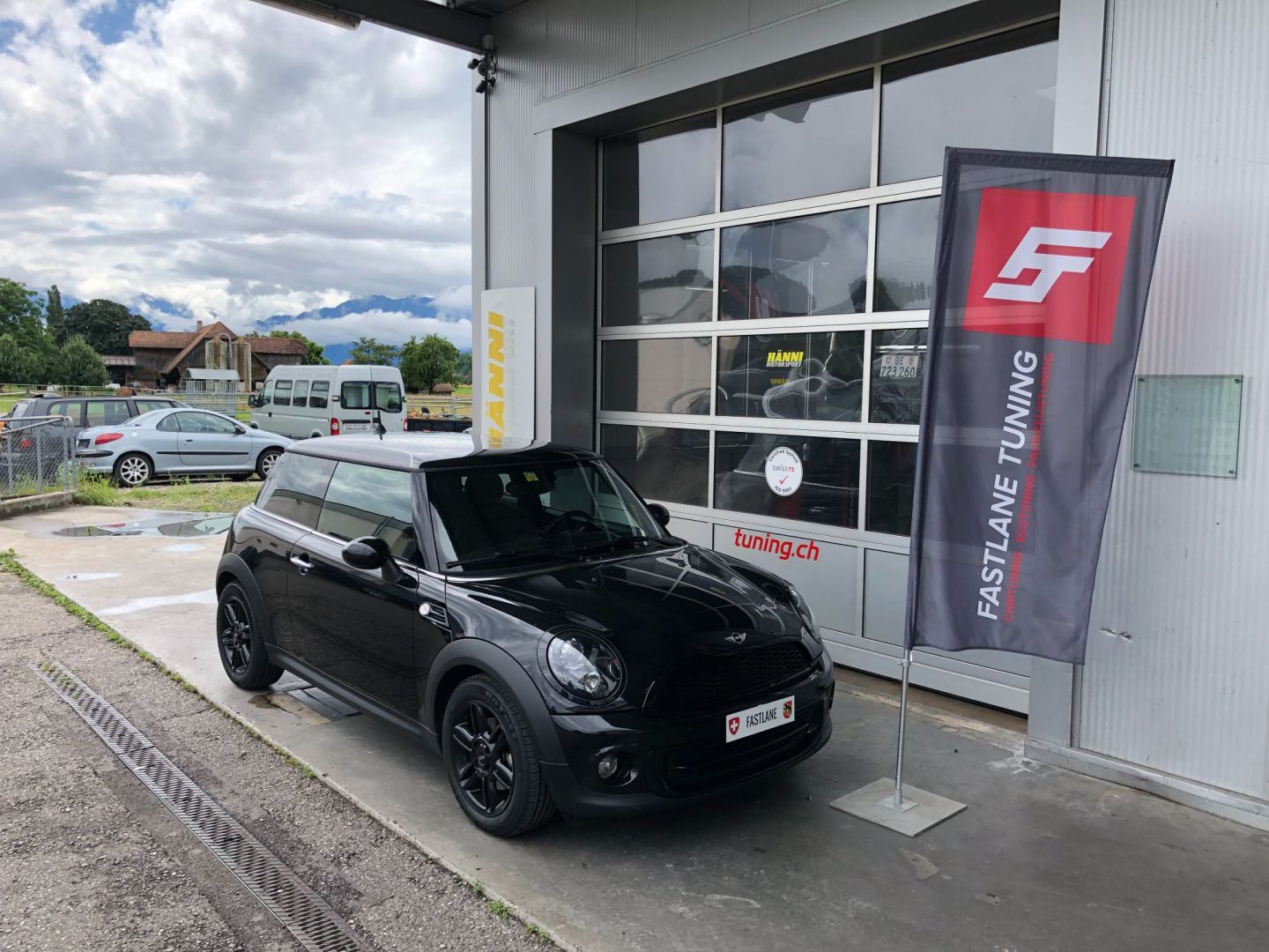 Ein kleiner schwarz getönter Mini One Hatch steht vor der Garage neben der Fastlane Tuning Schweiz Flagge