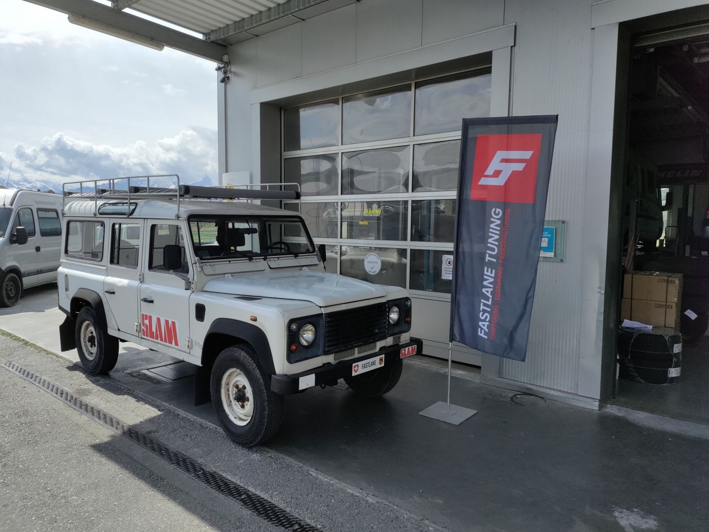 Ein weisser 110 Defender Td5 parkiert vor der Garage neben der Fastlane Tuning Schweiz Flagge