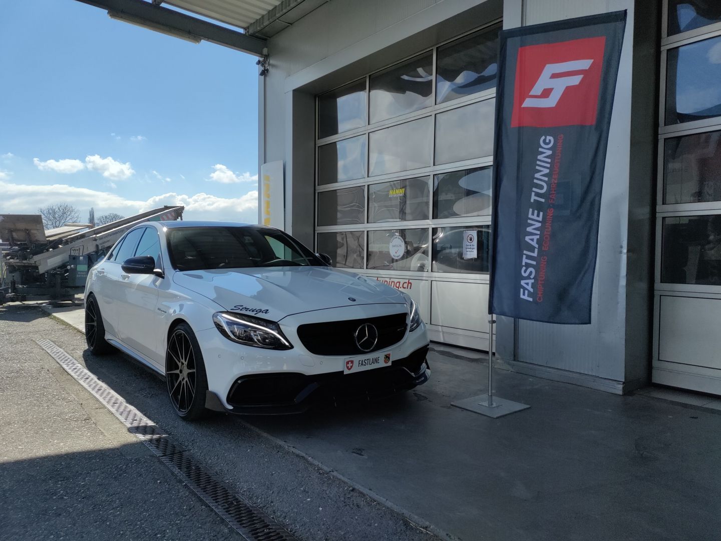 Ein weisser Mercedes Benz C63 AMG steht vor der Garage neben der Fastlane Tuning Schweiz Flagge