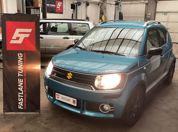 Ein aquablauer Suzuki Ignis 1.2 steht neben dem Banner von Fastlane Tuning Schweiz in der Garage