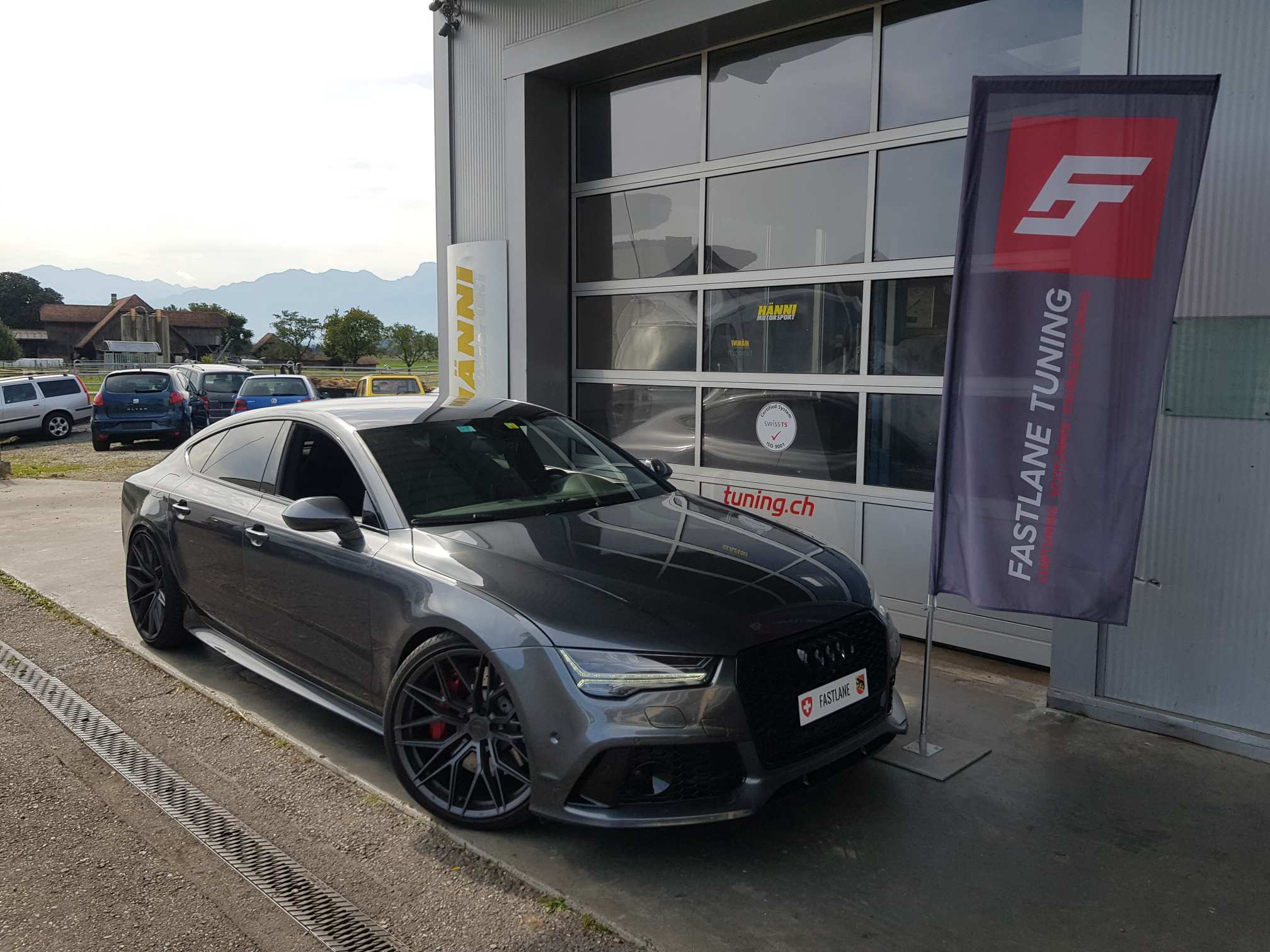 Ein dunkelgrauer Audi RS7 mit schwarzen Felgen steht vor der Garage neben der Fastlane Tuning Schweiz Flagge