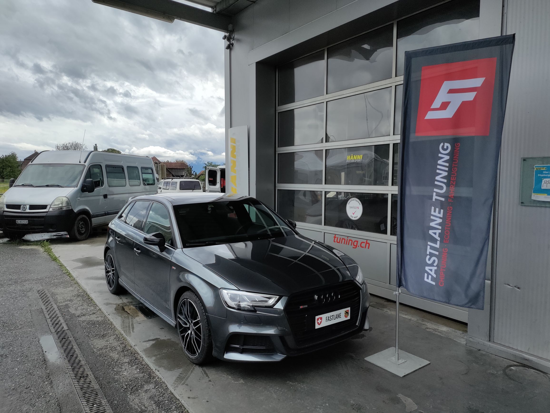 Ein anthrazitfarbener Audi A3 2.0 TFSI vor der Garage neben der Fastlane Tuning Schweiz Flagge