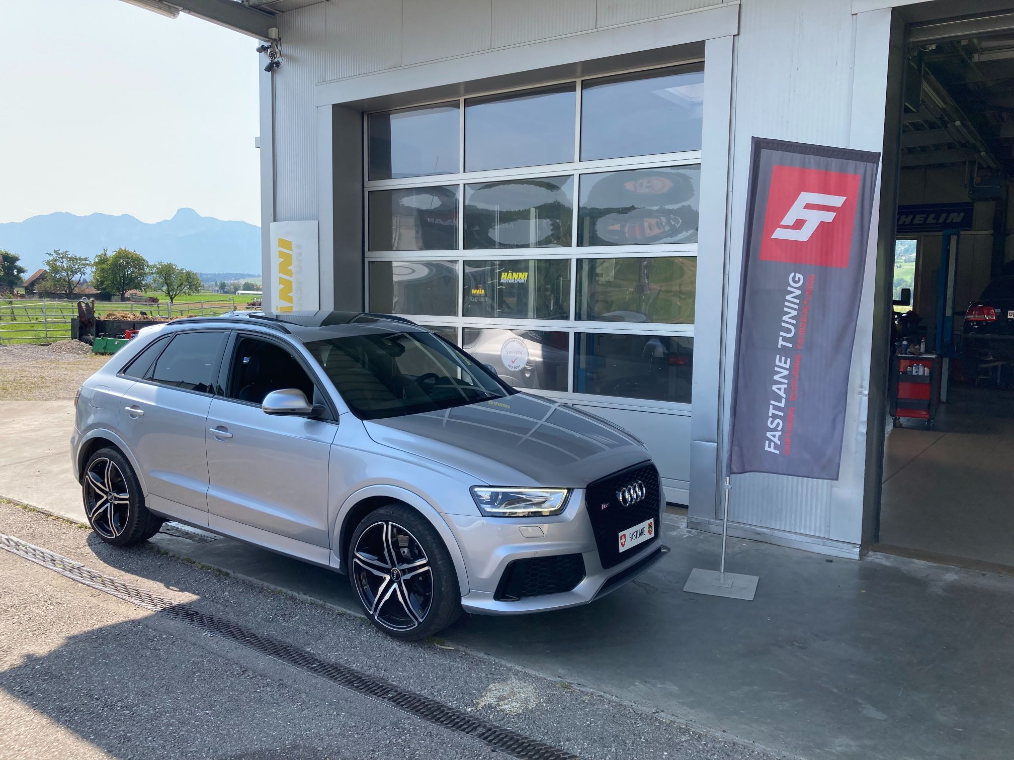 Ein silberfarbener Audi RSQ3 parkt vor der Garage neben der Fastlane Tuning Schweiz Flagge