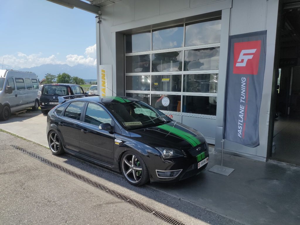 Ein schwarzer Ford Focus ST 225 mit grünem Rennstreifen über Motorhaube und Dach steht vor der Garage neben der Fastlane Tuning Schweiz Flagge