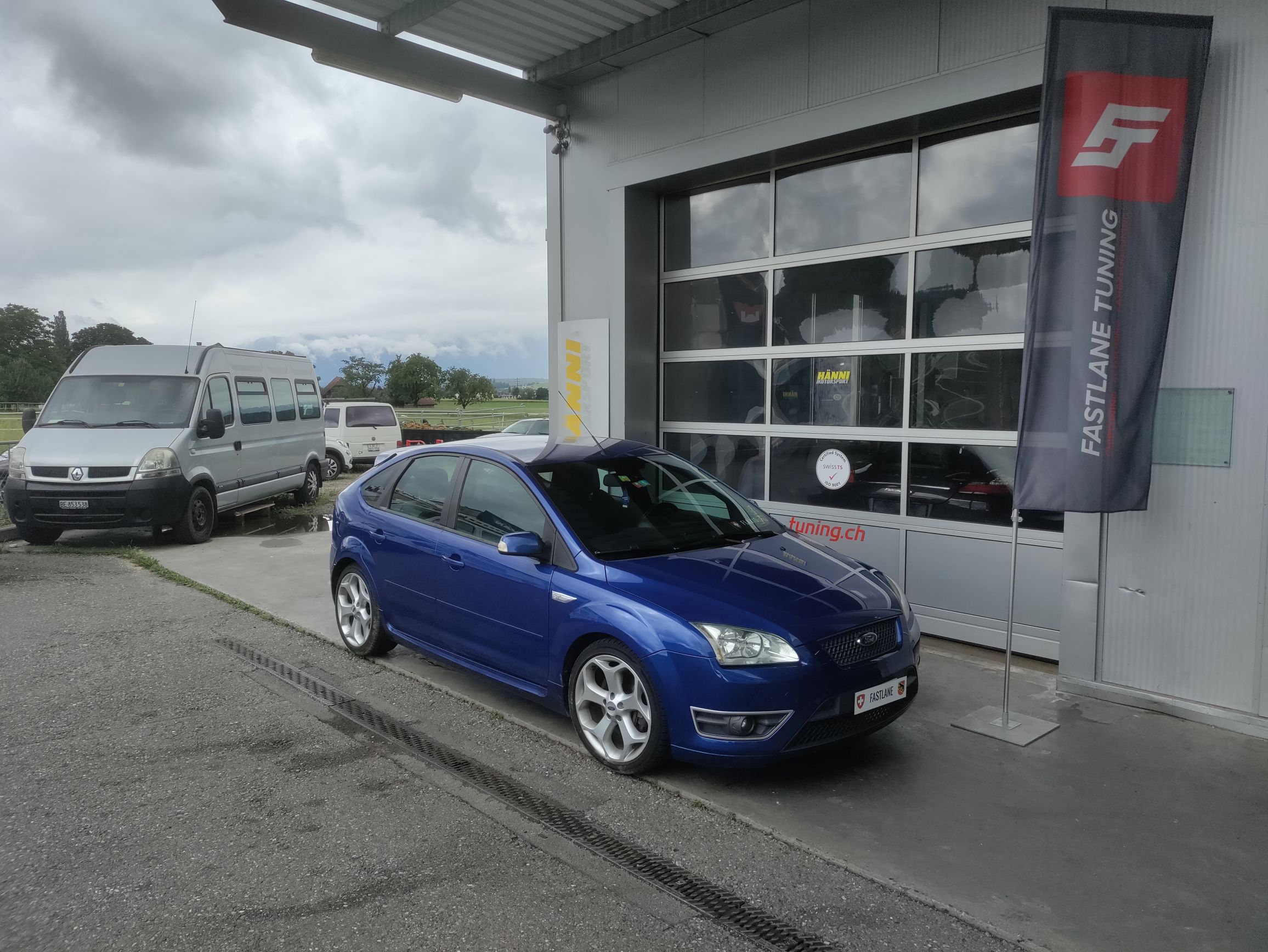 Ein blauer Ford Focus ST 225 mit Stage 1 Tuning steht vor der Garage neben der Fastlane Tuning Schweiz Flagge
