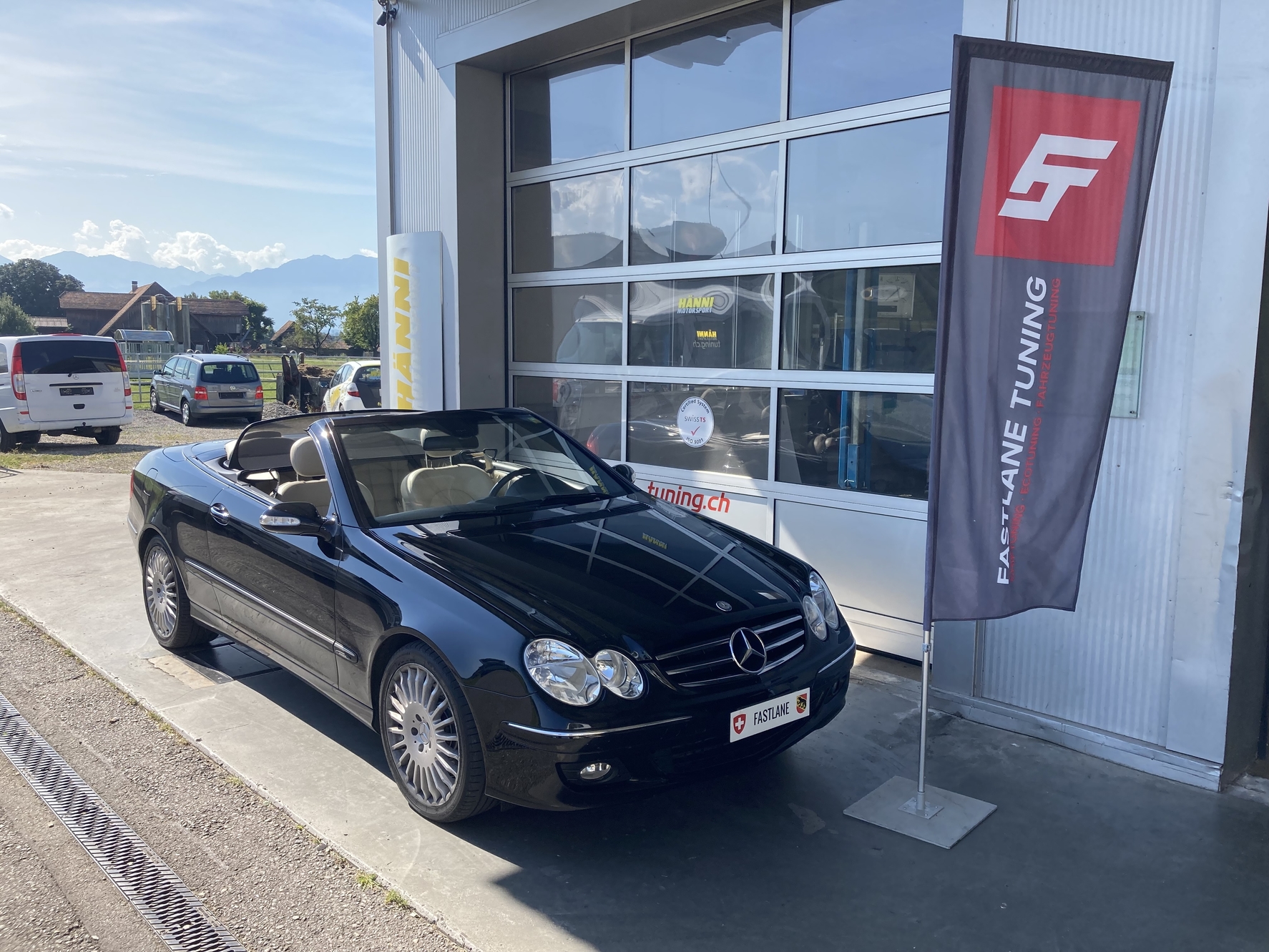 Ein schwarzes Cabrio Mercedes CLK 320 Diesel V6 steht vor der Garage neben der Fastlane Tuning Schweiz Flagge