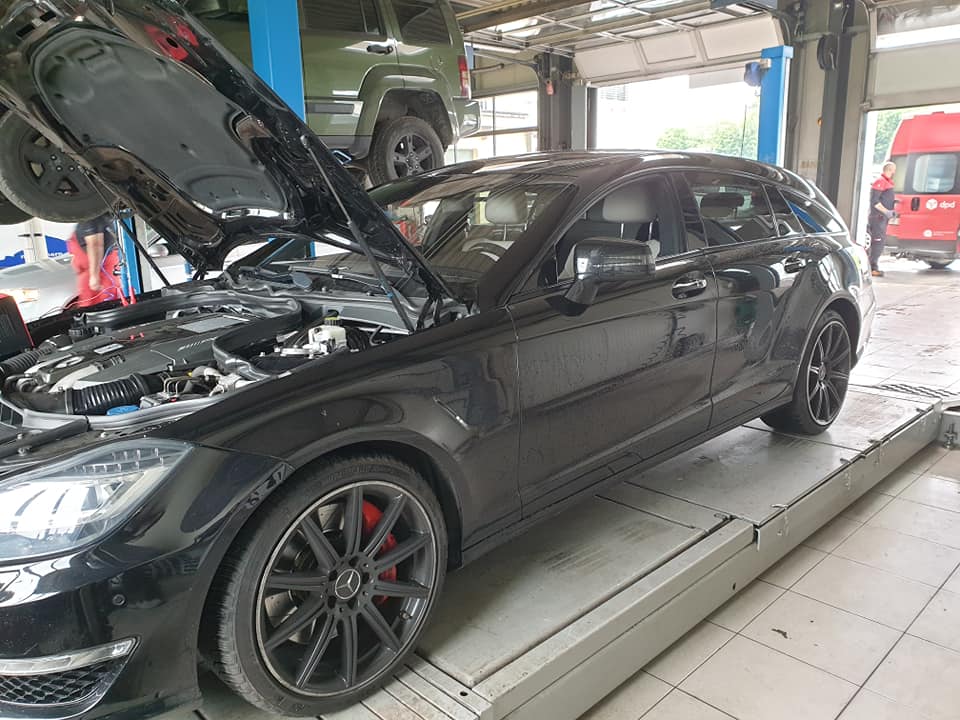 Ein schwarzer Mercedes Benz CLS 63 AMG steht auf einer Hebebühne in der Garage, wobei die Motorhaube geöffnet ist und den Blick auf den Motorraum freigibt. Er ist schwarz und hat schwarze Räder.