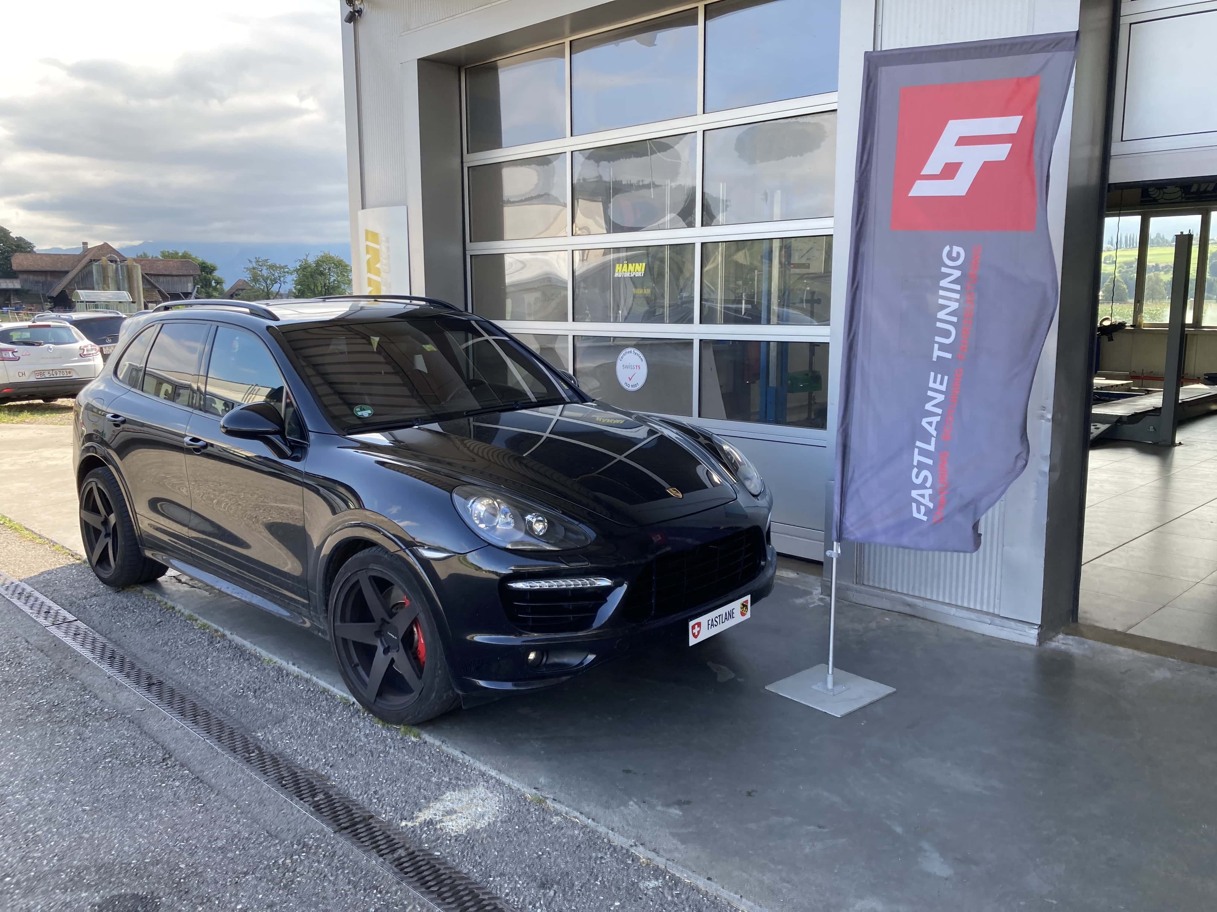 Ein schwarzer Porsche Cayenne Turbo steht vor der Garage neben der Fastlane Tuning Schweiz Flagge
