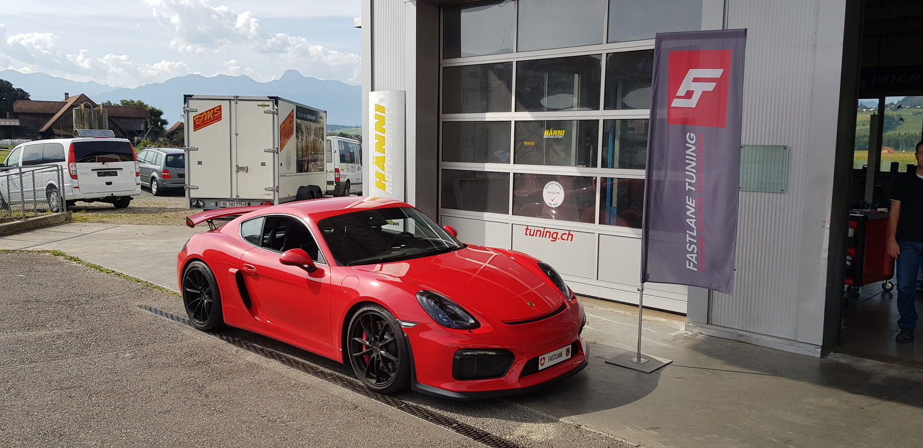 Ein roter Cayman GT4 steht vor der Garage neben der Fastlane Tuning Schweiz Flagge
