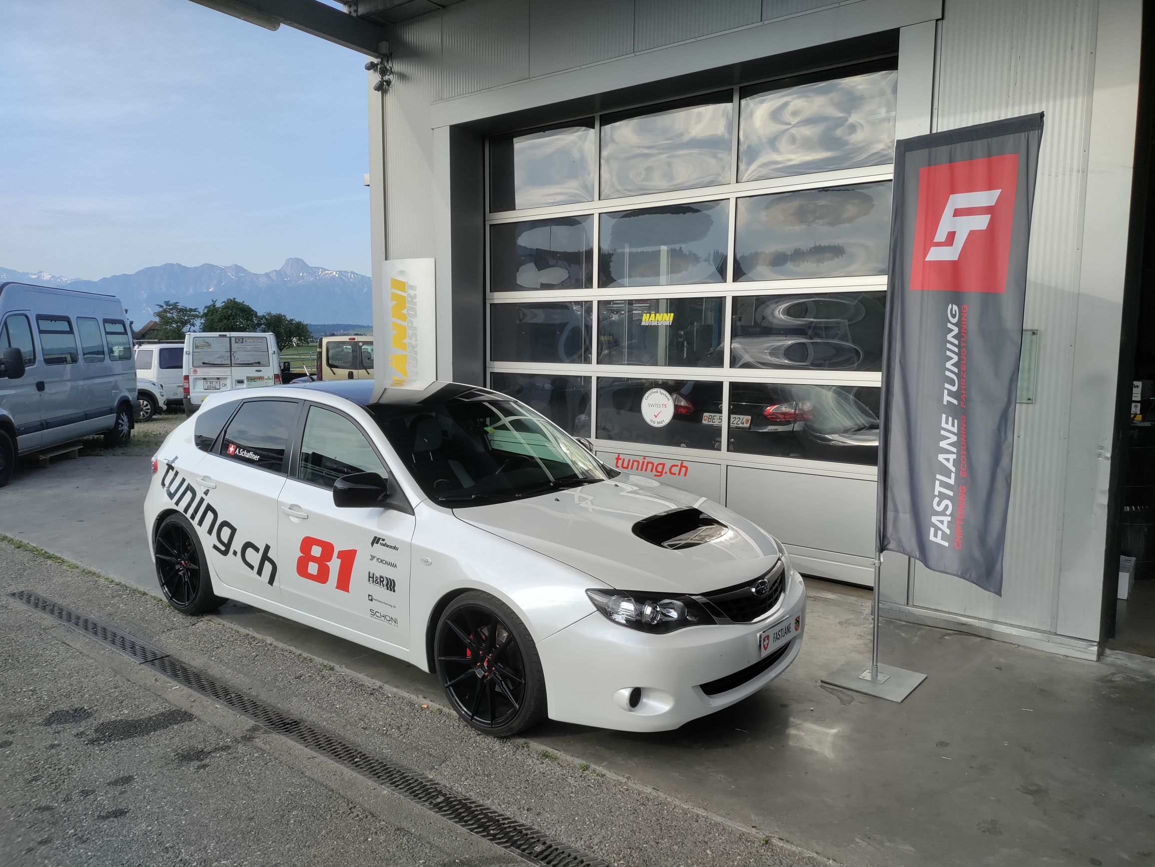 Ein weisser Subaru Impreza Diesel mit Rennaufklebern und einer Rennnummer parkiert vor der Garage neben der Fastlane Tuning Schweiz Flagge