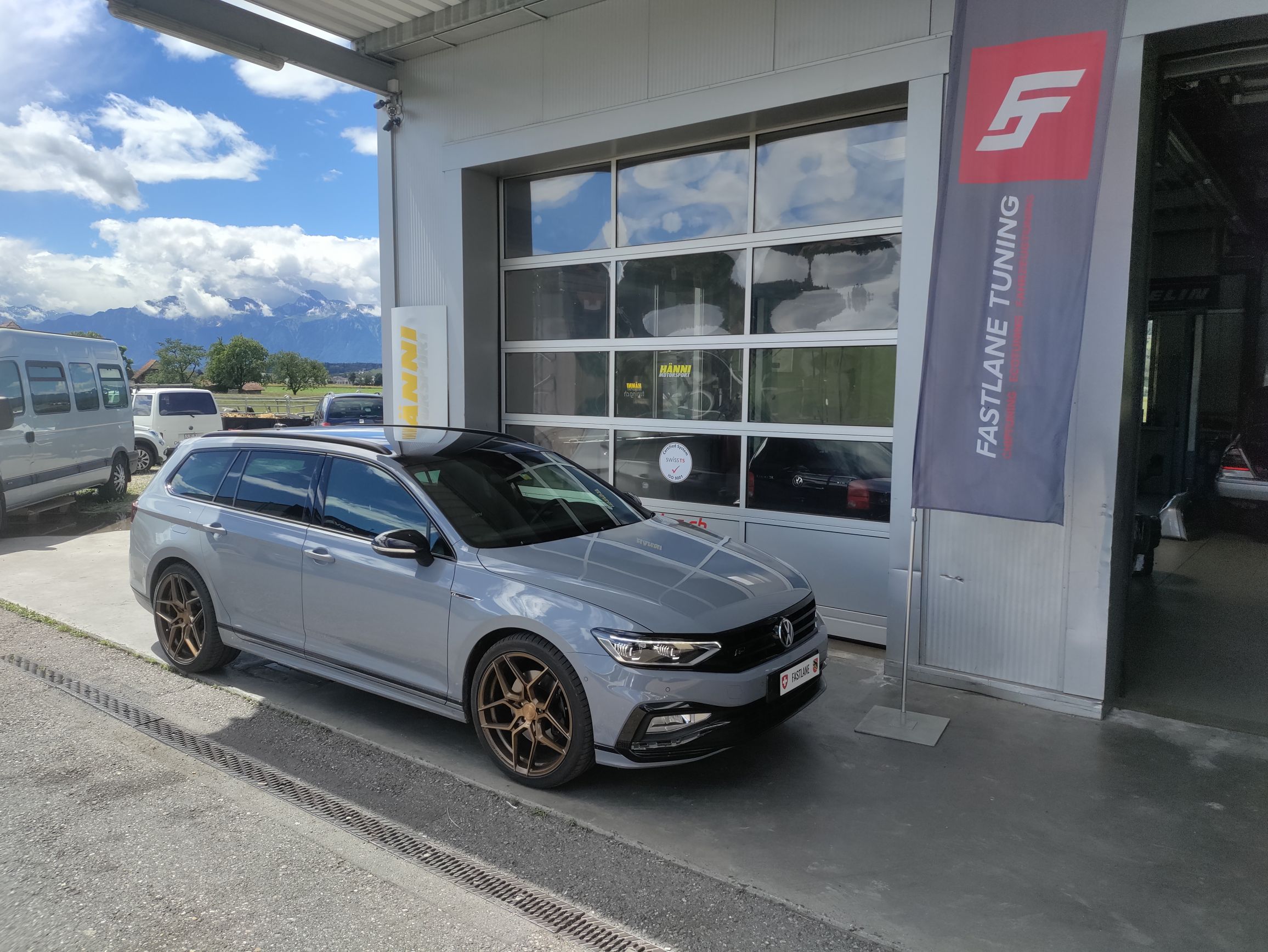 Ein mattgrauer Volkswagen Passat R Line steht vor der Garage neben der Fastlane Tuning Schweiz Flagge