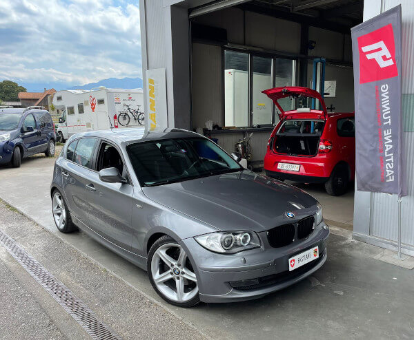 Ein grauer BMW 120i steht neben der Fastlane Tuning Schweiz Flagge.