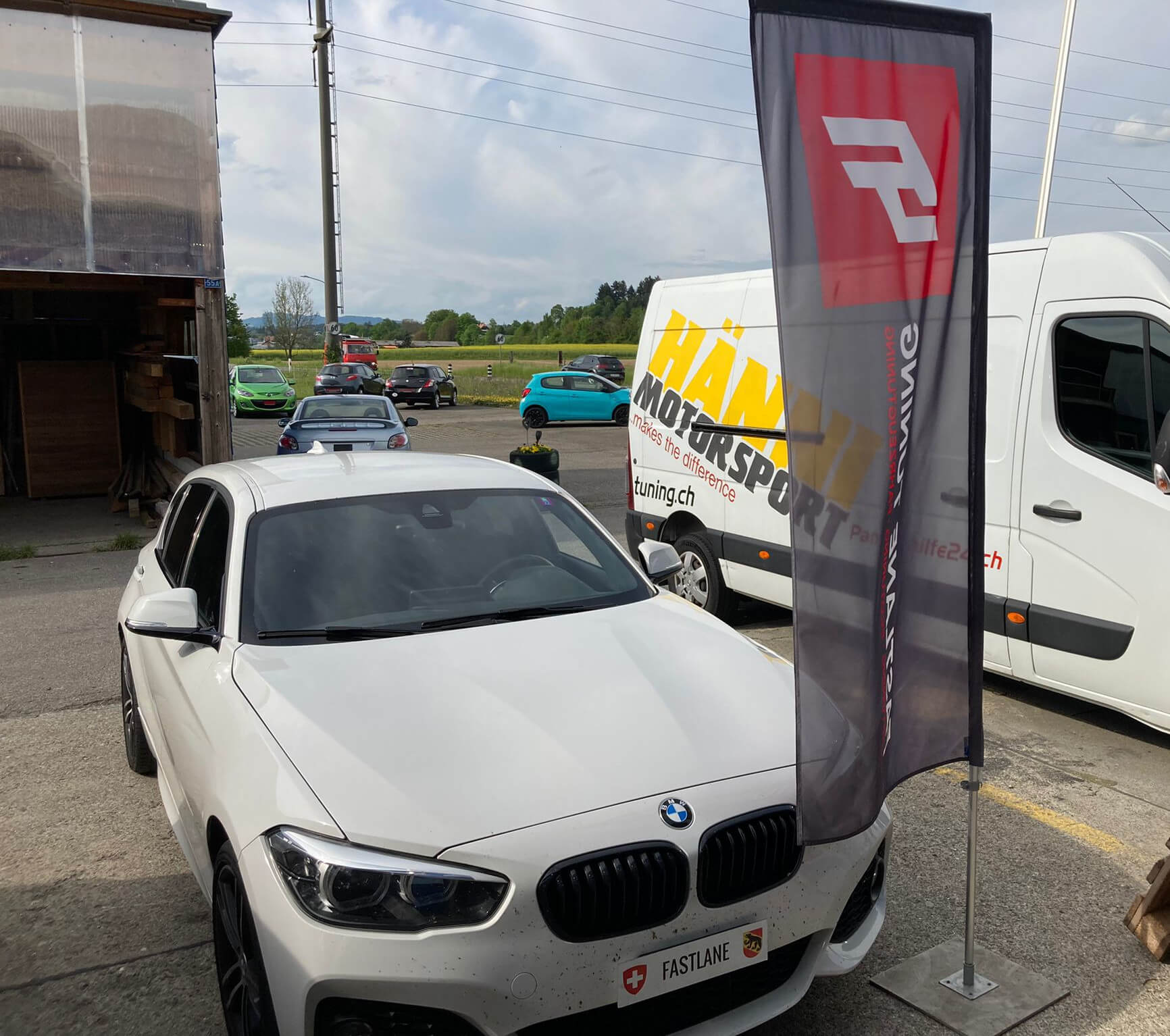 Ein weisser BMW 120i steht ausserhalb der Garage neben der Fastlane Tuning Schweiz Flagge und neben dem Hanni Motorsport Wagen.