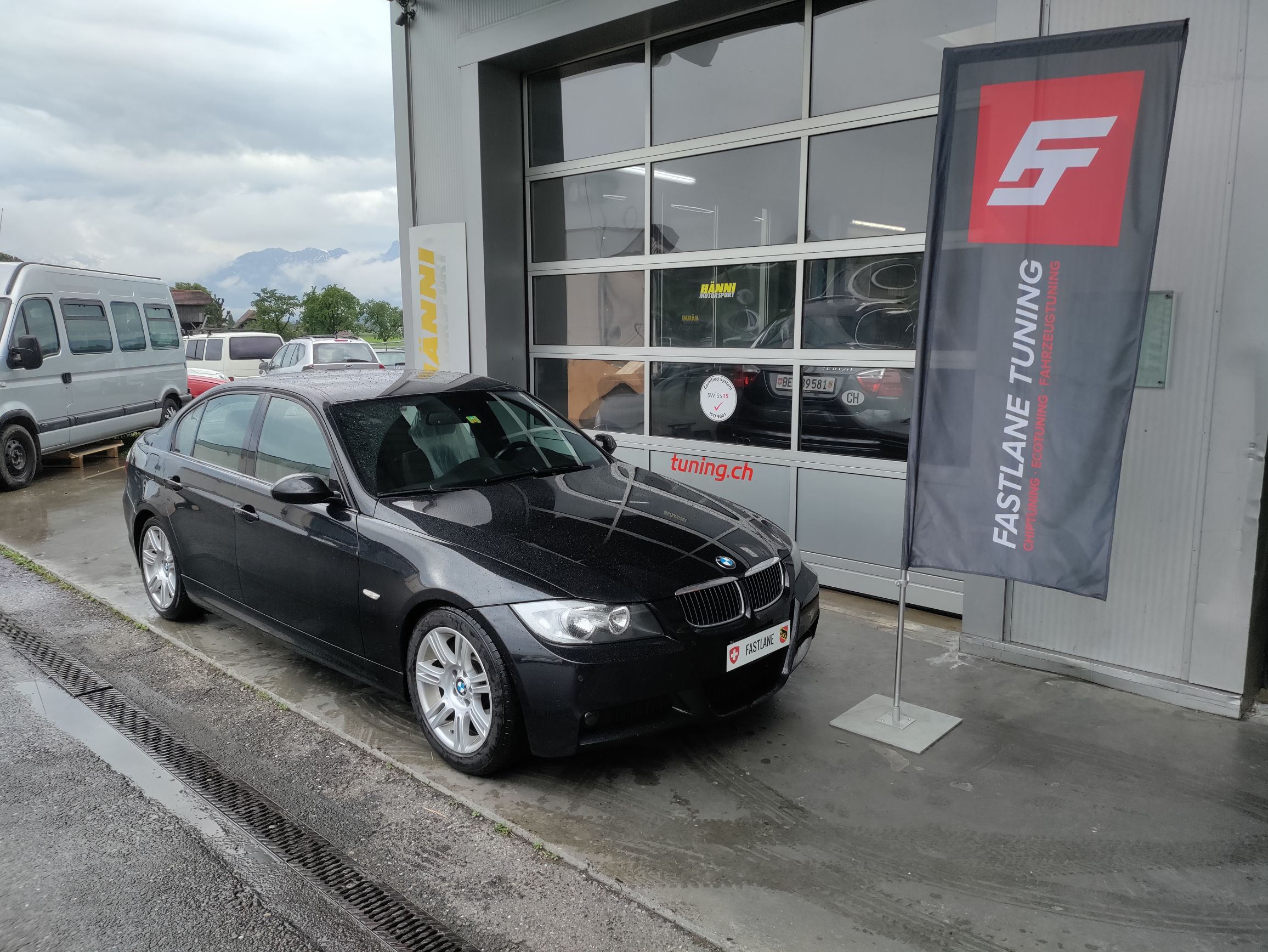 Ein schwarzer BMW E90 335 Diesel steht vor der Garage neben der Fastlane Tuning Schweiz Flagge.
