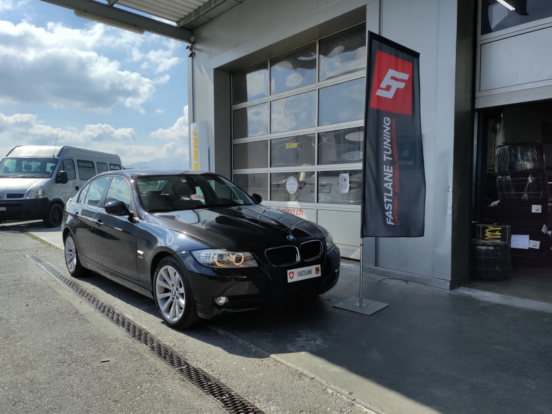 Ein schwarzer BMW 335i E Series mit N55-Motor steht vor der Garage neben der Fastlane Tuning Schweiz Flagge.