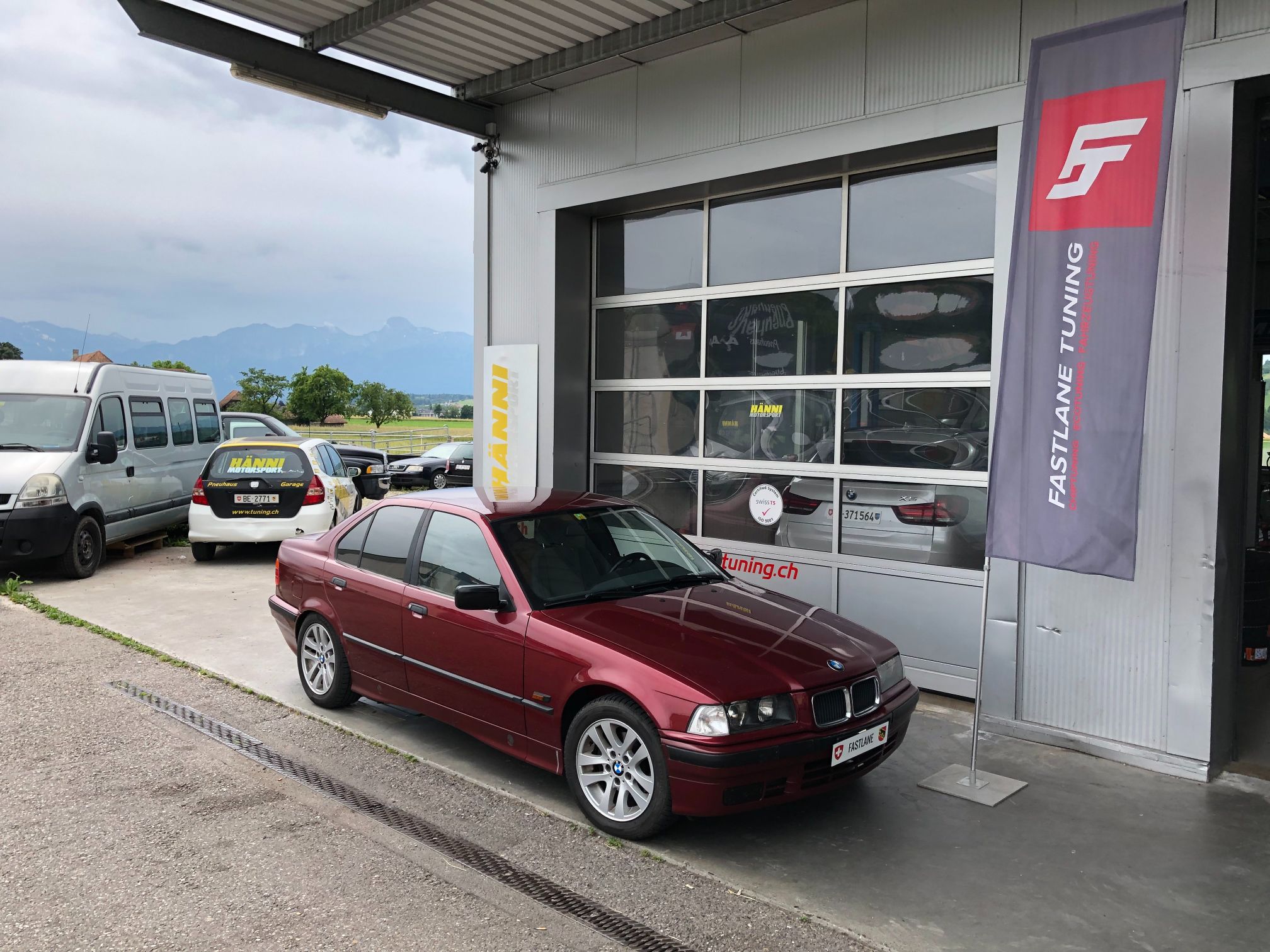 Ein dunkelroter BMW E36 323i mit silbernen Felgen steht vor der Garage neben der Fastlane Tuning Schweiz Flagge.