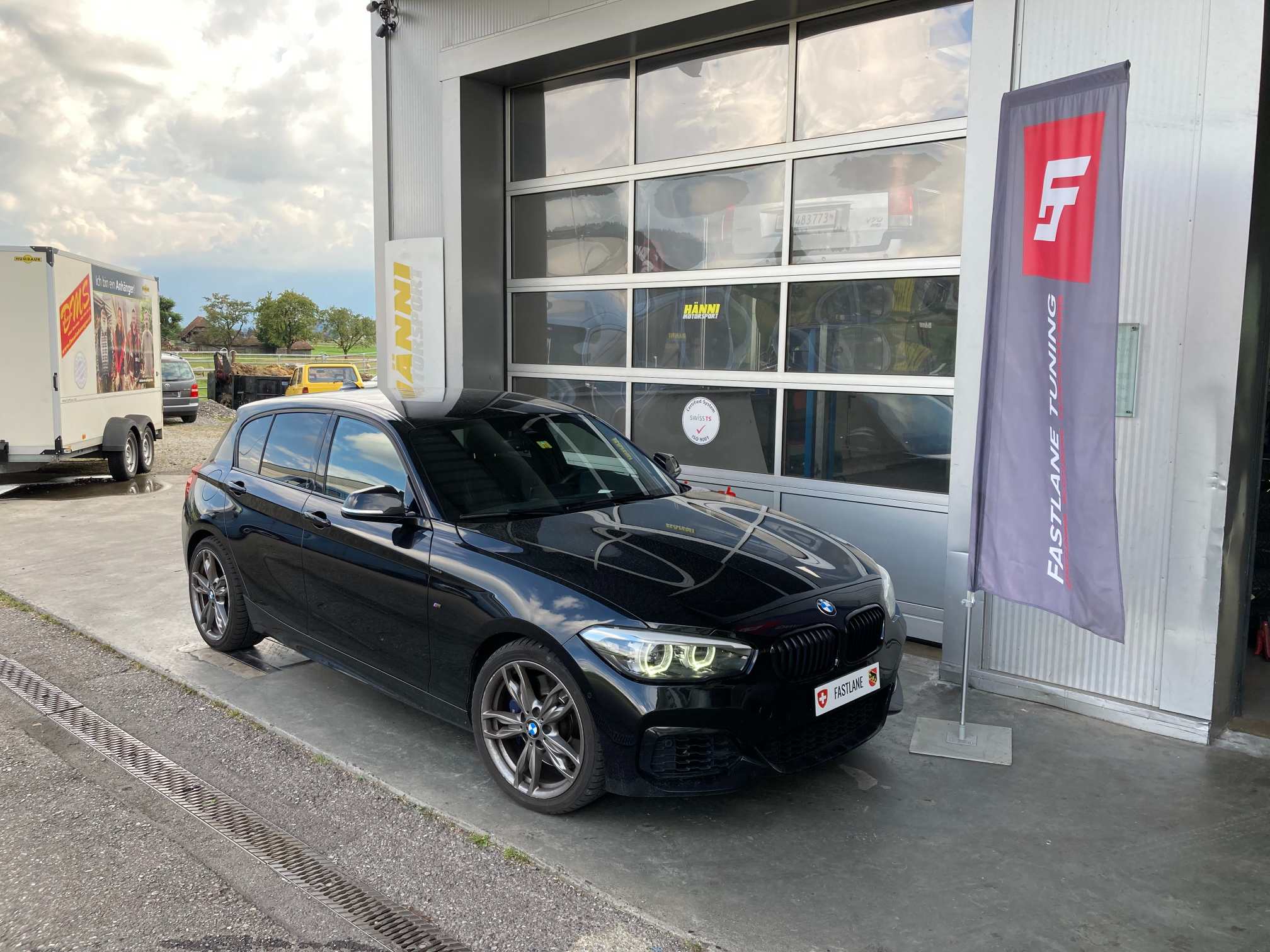 Ein schwarzer BMW M140i Shadow Edition parkt vor der Garage neben der Fastlane Tuning Schweiz Flagge.