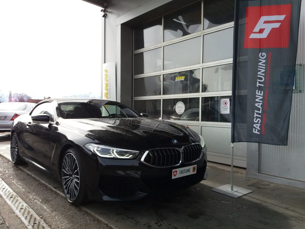 Ein schwarzes BMW M850i Cabriolet parkt vor der Garage neben der Fastlane Tuning Schweiz Flagge.