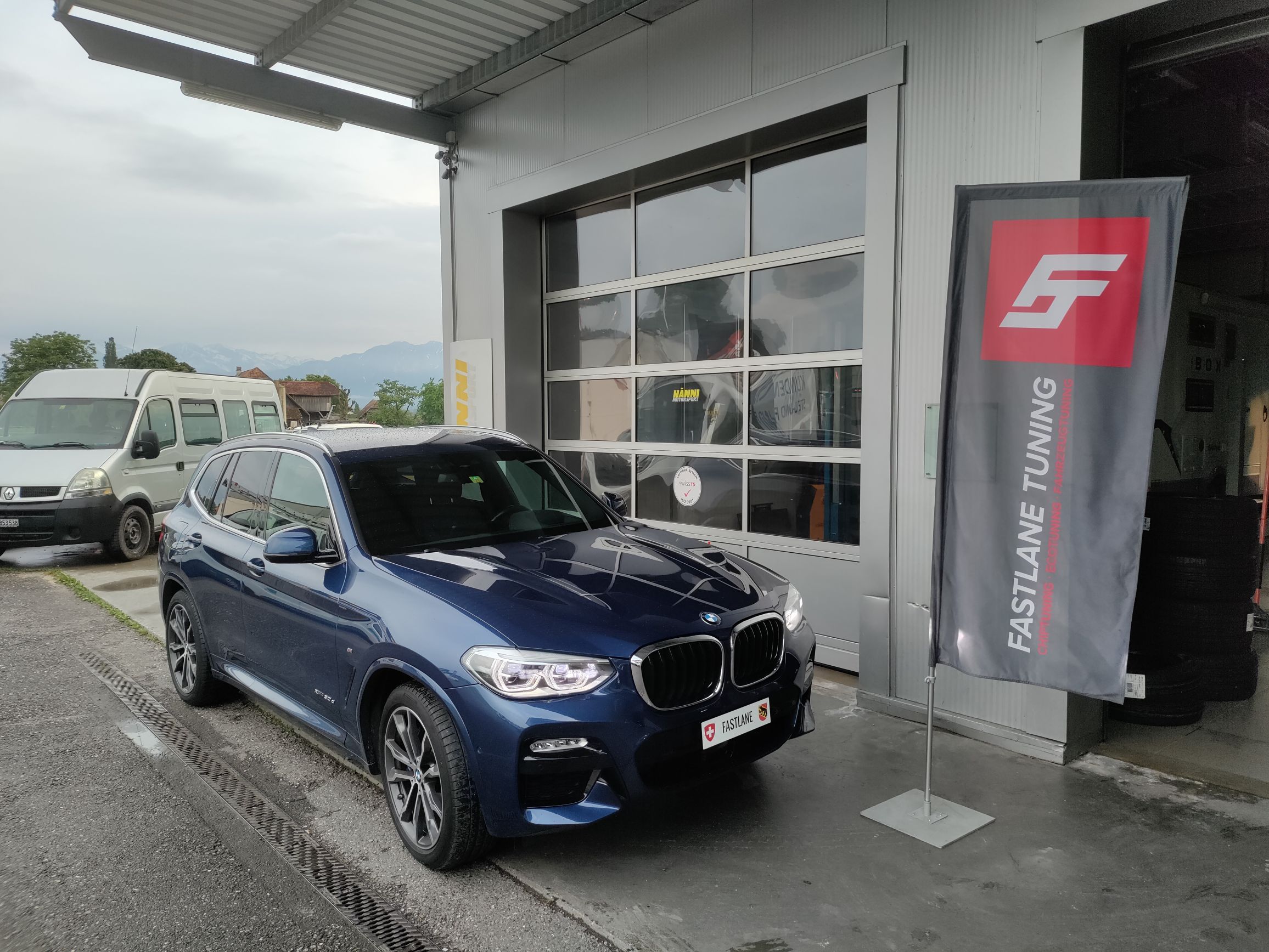 Ein dunkelblauer BMW X3 30d 2.0 Diesel steht vor der Garage neben der Fastlane Tuning Schweiz Flagge