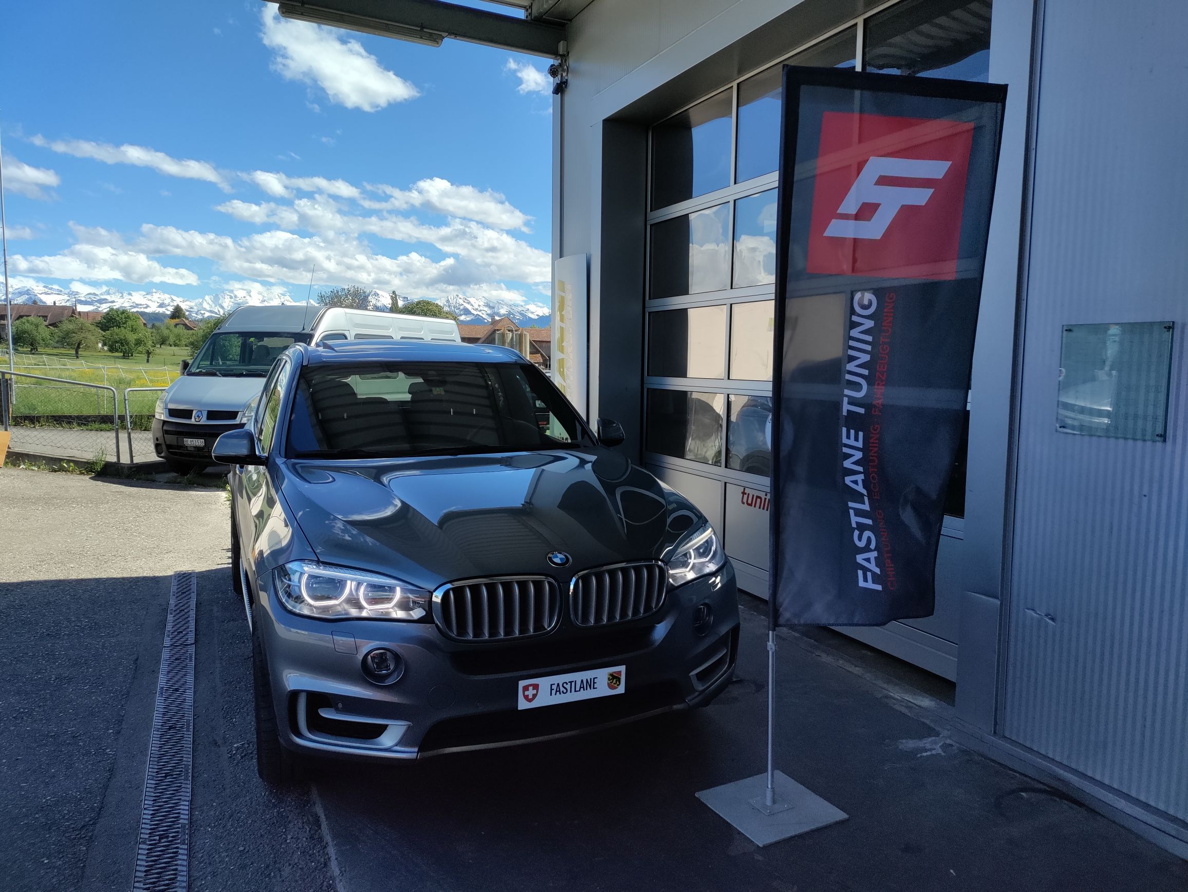 Ein silbergrauer BMW X5 50i V8 steht vor der Garage neben der Fastlane Tuning Schweiz Flagge