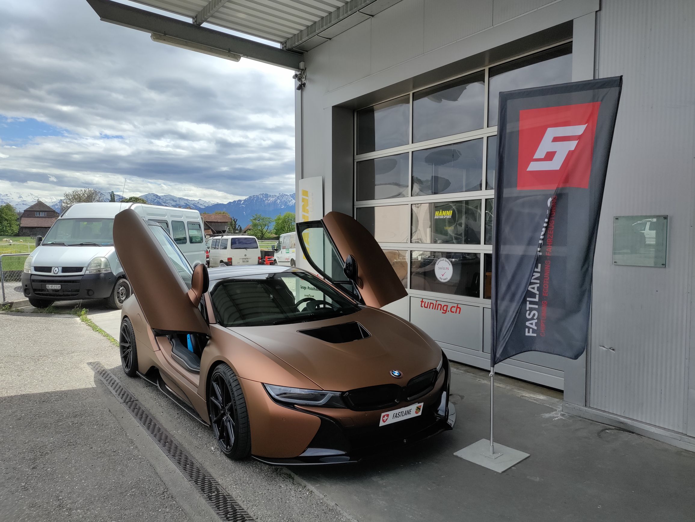 Ein brauner BMW i8 hybrid drive mit geöffneten Flügeltüren steht vor der Garage neben der Fastlane Tuning Schweiz Flagge.